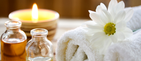 Rolled up towels with a white flower on them, next to spa massage oils and a brightly burning yellow candle.